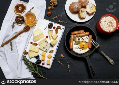 fried egg with sausages in a black round cast-iron frying pan, red mug with cocoa and marshmallow and cheese board with various kinds of cheese on a black wooden table