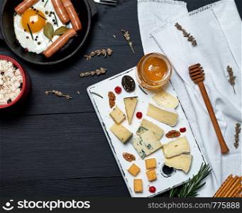 fried egg with sausages in a black round frying pan with a handle, a board with sliced different cheese and a cup of coffee, breakfast for one, top view