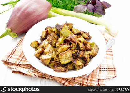 Fried eggplant with onion, egg and spices in a plate on a napkin, basil, leek and dill on white wooden board background