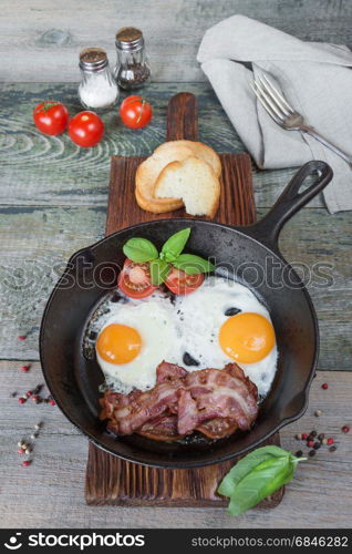 Fried eggs, strips of bacon, halved tomato and basil in a cast iron skillet on an old wooden table in rustic style