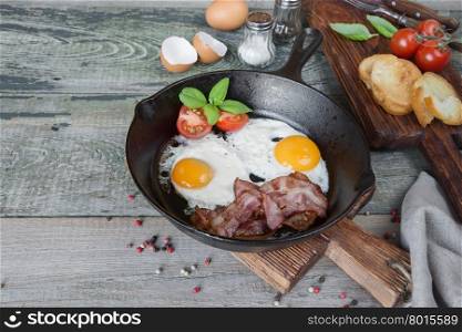 Fried eggs, strips of bacon, halved tomato and basil in a cast iron skillet on an old wooden table in rustic style