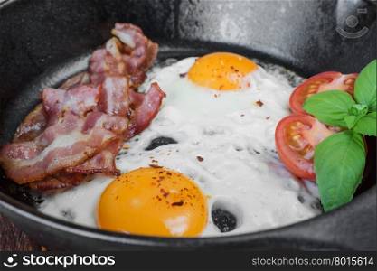 Fried eggs, strips of bacon, halved tomato and basil in a cast iron skillet close-up in rustic style