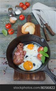 Fried eggs, strips of bacon, halved tomato and basil in a cast iron skillet on an old wooden table in rustic style