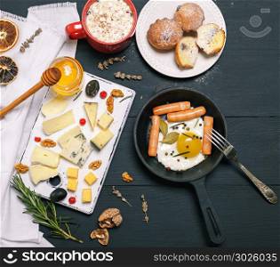 fried eggs with sausages in a black round frying pan and cheese board with a jar of honey on a black wooden background, top view, breakfast for one