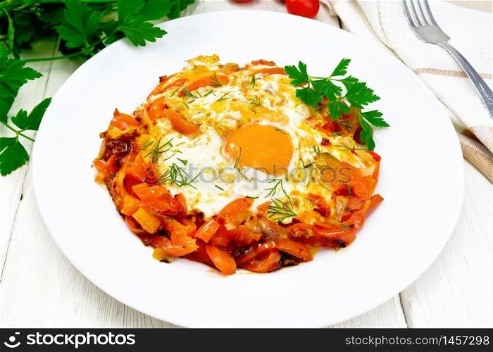 Fried eggs with tomatoes, sweet pepper, onions and herbs in a plate, napkin, parsley and fork on background of light wooden board