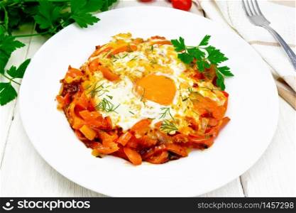 Fried eggs with tomatoes, sweet pepper, onions and herbs in a plate, napkin, parsley and fork on background of light wooden board