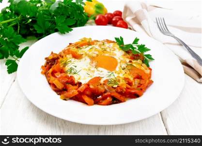 Fried eggs with tomatoes, sweet pepper, onions and herbs in a plate, napkin, parsley and fork on white wooden board background