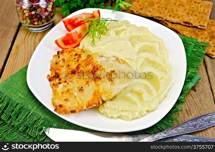 Fried fish with mashed potatoes and tomatoes on a plate, pepper, parsley, napkin, knife and fork on a wooden board
