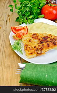 Fried fish with mashed potatoes on a plate, parsley, tomatoes, napkin, knife and fork on a wooden board