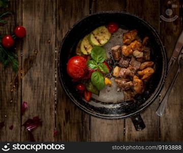 fried pieces of meat and vegetables on an old frying pan, view from above. wooden background. dish on a wooden surface