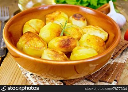 Fried potatoes with rosemary in a ceramic pan on a napkin, garlic, parsley, vegetable oil, fork on a wooden boards background