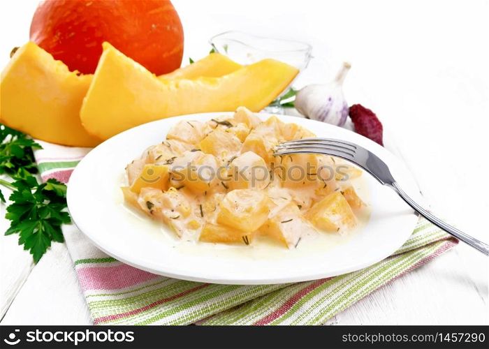 Fried pumpkin with garlic, spices and herbs in sour cream sauce in a plate on towel, parsley and dill, fork on wooden board background