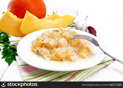 Fried pumpkin with garlic, spices and herbs in sour cream sauce in a plate on towel, parsley and dill, fork on wooden board background