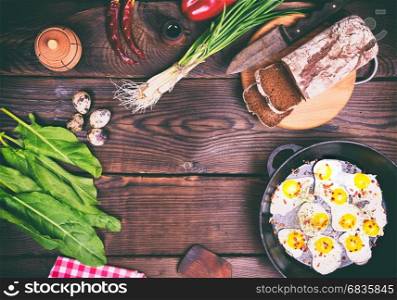 Fried quail eggs in a black cast-iron frying pan, near rye bread on a cutting board and fresh vegetables, top view, empty space in the middle, vintage toning