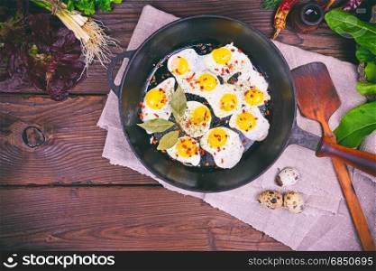 Fried quail eggs in a black cast-iron frying pan on a brown wooden background, top view