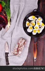 Fried quail eggs in a cast-iron black frying pan with a wooden handle on the table, next to a whole loaf of rye bread, top view