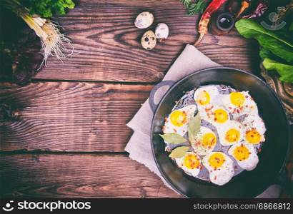 Fried quail eggs in a cast-iron frying pan and fresh vegetables on a brown wooden table, empty space in the middle