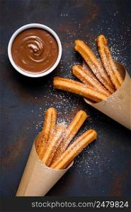 Fried traditional churro sticks with sugar powder cinnamon and bowl of chocolate dip on dark table top view