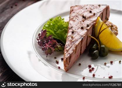 Fried Tuna Steak on Round White Plate with Seasoning and Fresh Green Salad with Lemon and capers on Wooden Table.