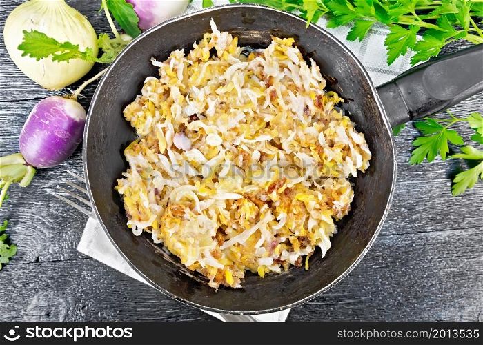 Fried turnips with onions in a frying pan on a towel on wooden board background from above