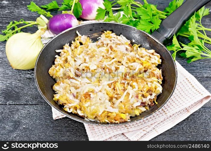 Fried turnips with onions in a frying pan on a towel on black wooden board background