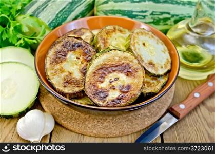 Fried zucchini in a ceramic pot, garlic, fresh zucchini, parsley, knife, oil in a bottle on a wooden boards background
