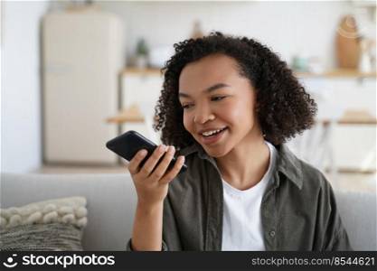Friendly african american young girl sit on couch at home hold smartphone leave voice message. Modern biracial female chat with friends use loudspeaker virtual assistant search information on internet. Friendly african american young girl hold smartphone leave voice message or use virtual assistant