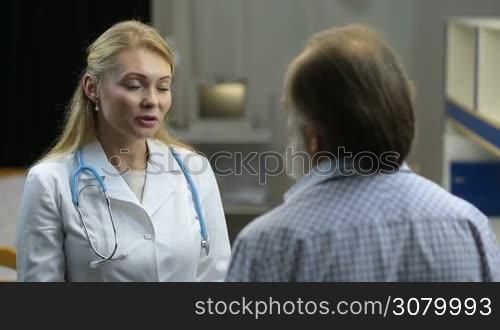 Friendly attractive female physician talking to ill stressed senior male patient and giving treatment advice in clinic. Cheerful woman doctar encouraging elderly man and touching patient&acute;s shoulder to show support and empathy after medical exam.