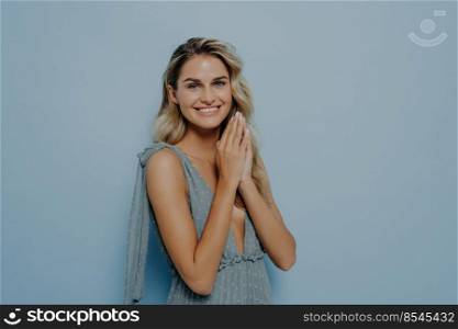 Friendly beautiful young blonde woman wearing romantic summer dress with trustworthy angel stance, holding hands in praying gesture, smiling and asking for help, standing alone next to blue background. Dreamy young blonde woman with hands pressed together under chin