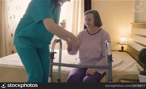 Friendly caregiver helping senior woman getting up from bed and walk with a walker. Home or hospice nursing and assistance concept.