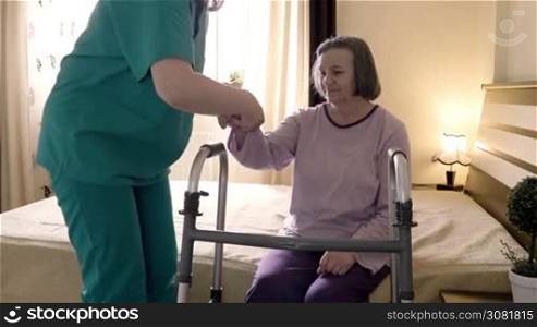 Friendly caregiver helping senior woman getting up from bed and walk with a walker. Home or hospice nursing and assistance concept. Slow motion