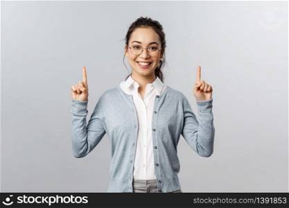 Friendly cheerful asian girl in glasses, showing product or company banner, pointing fingers up inviting check-up event news, info about advertisement, smiling adorable at camera, grey background.. Friendly cheerful asian girl in glasses, showing product or company banner, pointing fingers up inviting check-up event news, info about advertisement, smiling adorable at camera, grey background