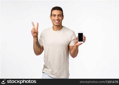 Friendly, cheerful handsome man in casual outfit, holding smartphone with blank screen, showing peace or victory sign, smiling, advertise site or app, recommend download mobile application.. Friendly, cheerful handsome man in casual outfit, holding smartphone with blank screen, showing peace or victory sign, smiling, advertise site or app, recommend download mobile application