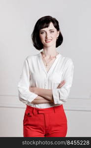 Friendly confident young woman with folded arms smiling at the camera. brunette Girl with make up, short hair wearing in white shirt and red pants on white wall background.. Friendly confident young woman with folded arms smiling at the camera. brunette Girl with make up, short hair wearing in white shirt and red pants on white wall background