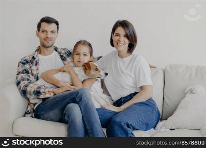 Friendly family pose together at sofa, enjoys domestic atmosphere. Father, mother, their little daughter and pedigree dog spend weekend at home, pose in living room, have happy face expressions