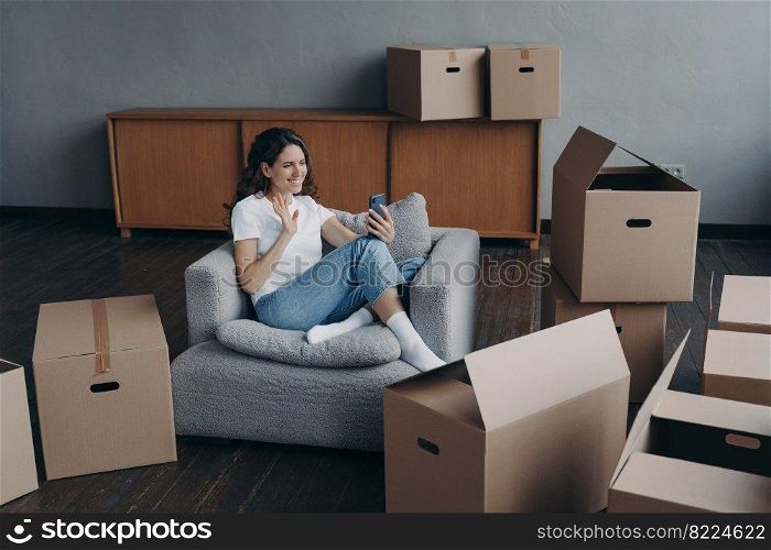 Friendly female blogger homeowner greeting followers in social networks, or chatting by video call, waving hand to camera, holding smartphone, sitting with cardboard boxes for relocation on moving day. Girl blogger greeting followers in social networks, sitting with boxes for relocation on moving day