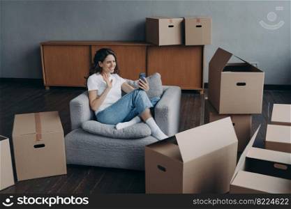 Friendly female blogger homeowner greeting followers in social networks, or chatting by video call, waving hand to camera, holding smartphone, sitting with cardboard boxes for relocation on moving day. Girl blogger greeting followers in social networks, sitting with boxes for relocation on moving day