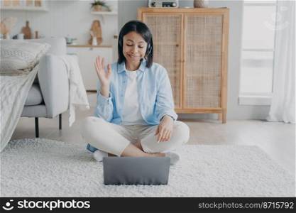 Friendly female coach in headset greeting client while online consultation on laptop by video call at home. Smiling woman in headphones conducts webinar welcomes students. Distant education, elearning. Female coach in headset greeting client while online consultation on laptop by video call at home
