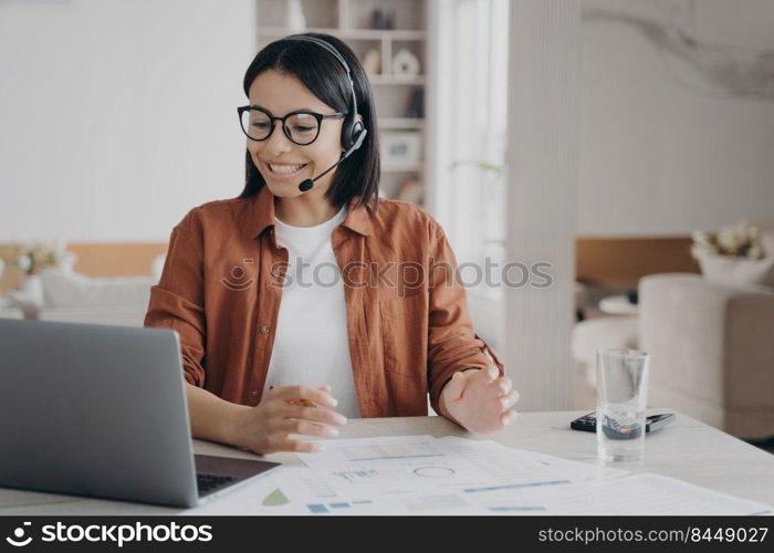 Friendly female coach wearing glasses and headset conducts webinar on laptop. Smiling businesswoman in headphones with microphone having video call, counseling client online looking at computer screen. Female coach in glasses, headset conducts online lesson on laptop. E-learning, distance education