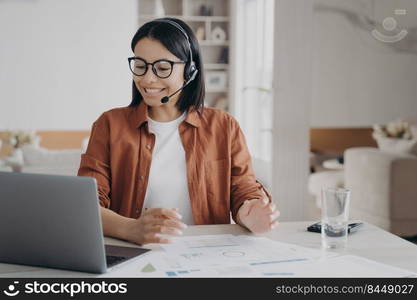 Friendly female coach wearing glasses and headset conducts webinar on laptop. Smiling businesswoman in headphones with microphone having video call, counseling client online looking at computer screen. Female coach in glasses, headset conducts online lesson on laptop. E-learning, distance education