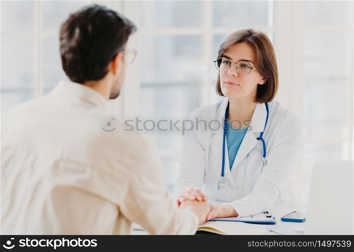 Friendly female doctor tries to support patient, holds his hands, gives useful consultation and explains medical information, makes diagnostic examining, pose in hospital room. Heathcare, assistance