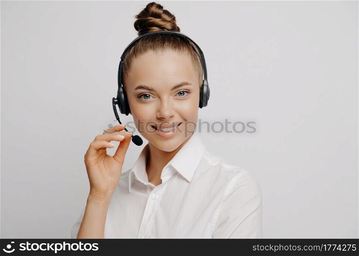 Friendly female marketing company agent with charmful smile in white shirt with black headset happy after finishing helping another satisfied customer, standing in front of light background. Friendly female marketing company agent with headset