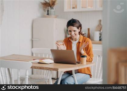Friendly lady has business video call by computer at home. Caucasian young woman in orange shirt is remote worker sitting at table at kitchen. Successful entrepreneur. Freelance distance work concept.. Friendly lady has business video call by computer at home. Freelance and distance work concept.