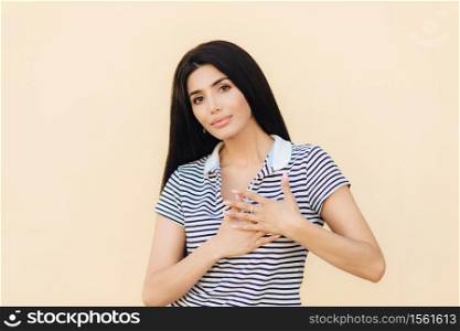 Friendly looking brunette female keeps hands on heart as expresses her gratitude, has serious expression, wears casual clothes, isolated over studio background. People, body language concept