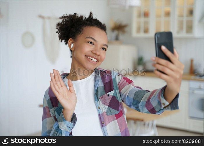 Friendly mixed race girl blogger holding phone, talking by video call, waving hand hello, greeting friends. Smiling young female in wireless earphone welcoming, chatting online by phone at home.. Friendly mixed race girl blogger holding phone, talking by video call, waving hand hello, greeting