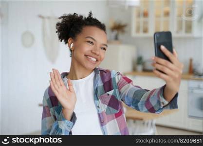 Friendly mixed race girl blogger holding phone, talking by video call, waving hand hello, greeting friends. Smiling young female in wireless earphone welcoming, chatting online by phone at home.. Friendly mixed race girl blogger holding phone, talking by video call, waving hand hello, greeting