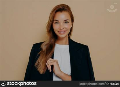 Friendly smiling brunette businesswoman with loose long hair, wears formal black jacket over white blouse, poses against beige background with blank space, happy to meet with business partners. Friendly smiling brunette businesswoman with loose long hair