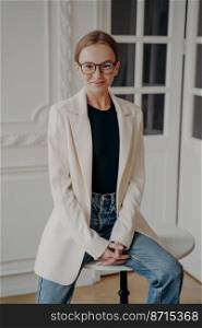 Friendly smiling businesswoman in glasses looking at camera, sitting on little round table. Confident female student wearing stylish casual clothing, white suit jacket and blue jeans, posing indoors.. Friendly smiling businesswoman in stylish glasses looking at camera, sitting on little round table
