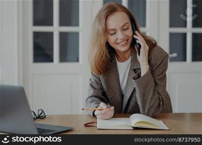 Friendly smiling female employee answers mobile call at desk with laptop. Businesswoman talking on phone enjoying pleasant conversation with colleague, consulting client making notes in notebook.. Female employee answers call at desk with laptop, talking on phone, consulting client, making notes