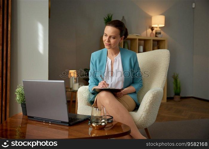Friendly smiling woman psychologist looking at laptop computer screen listening to patent and writing notes in clipboard. Psychotherapist at remote video conference meeting for therapy session. Friendly smiling woman psychologist looking at laptop computer screen